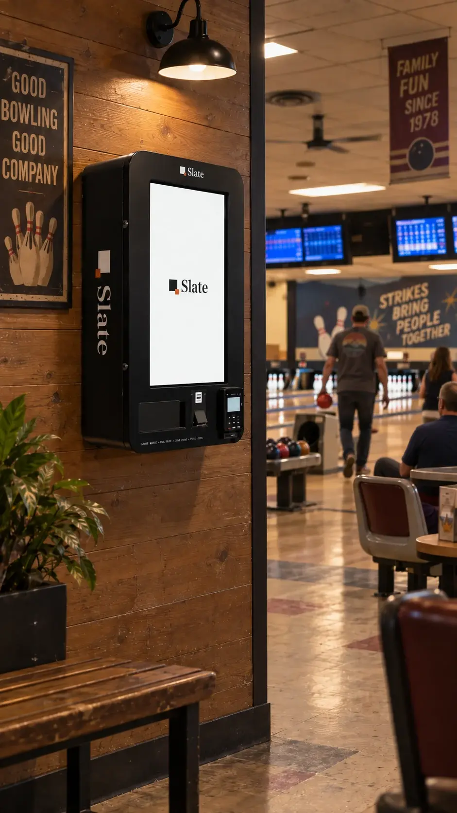 The kiosk on a plank wall at a bowling alley, a lamp above it, a plant and a bench to the left, and the lanes, scoreboards and bowlers through the opening to the right.