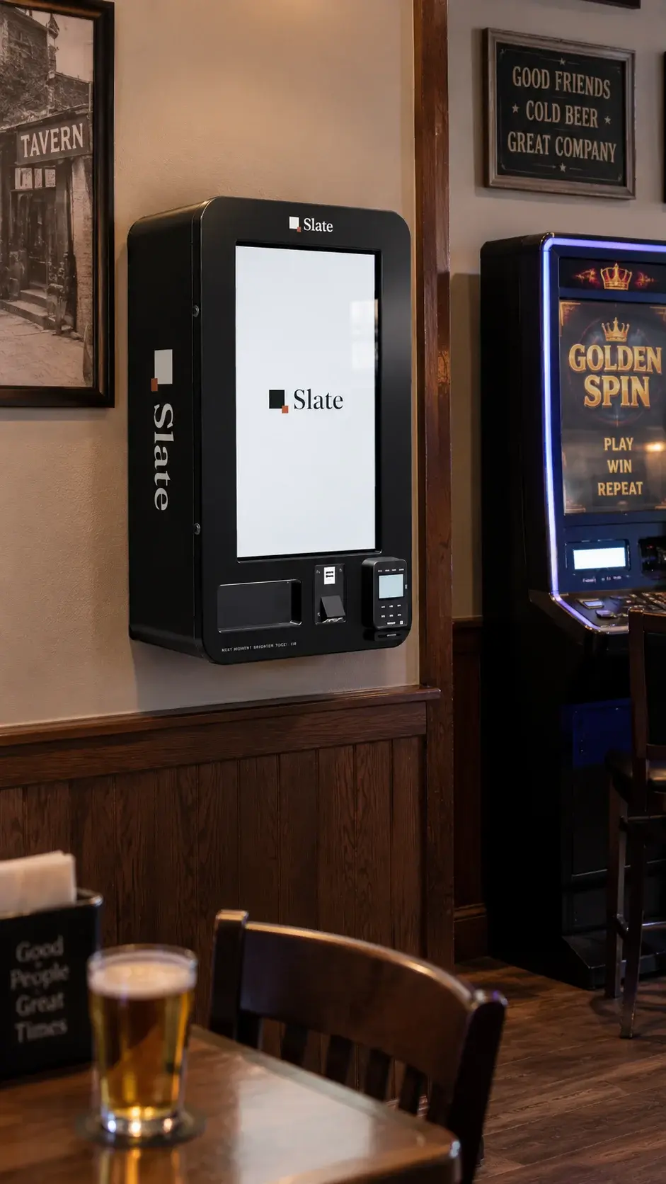 The kiosk on a plaster wall above dark wainscot in a tavern, a video gaming terminal to its right, framed pictures around it and a table with a pint in the foreground.